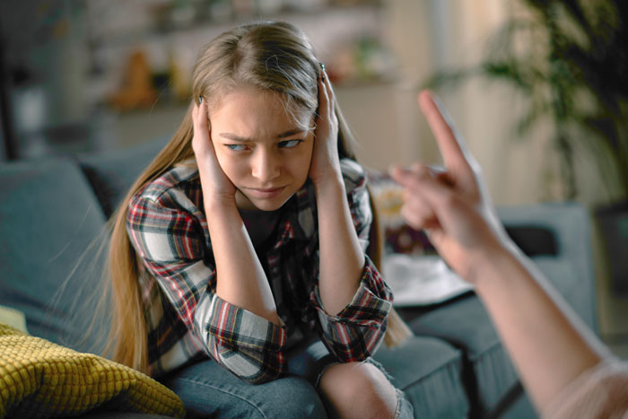 Teen girl covering ears while sitting on couch during a tense conversation about dad pulling funding for her trip.