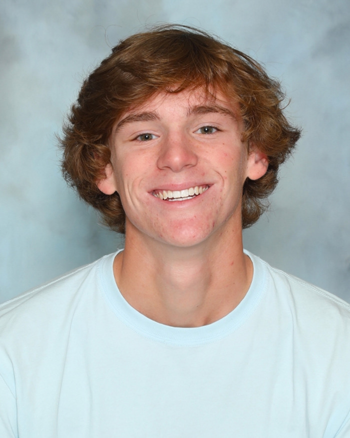 Teen golfer smiling in a light blue shirt in a school photo with a blurred gray background.