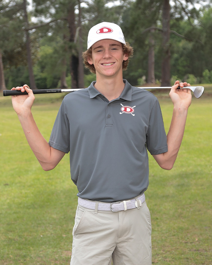 Teen golfer smiling outdoors in team uniform holding golf club behind his shoulders on a golf course.
