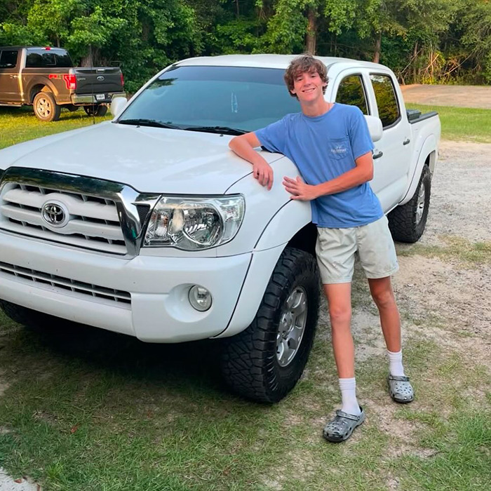Teen golfer in blue shirt and shorts leaning on a white pickup truck outdoors with trees and grass in the background.