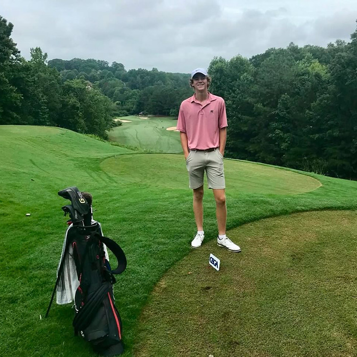 Teen golfer standing on a golf course near a golf bag, surrounded by trees and cloudy skies.