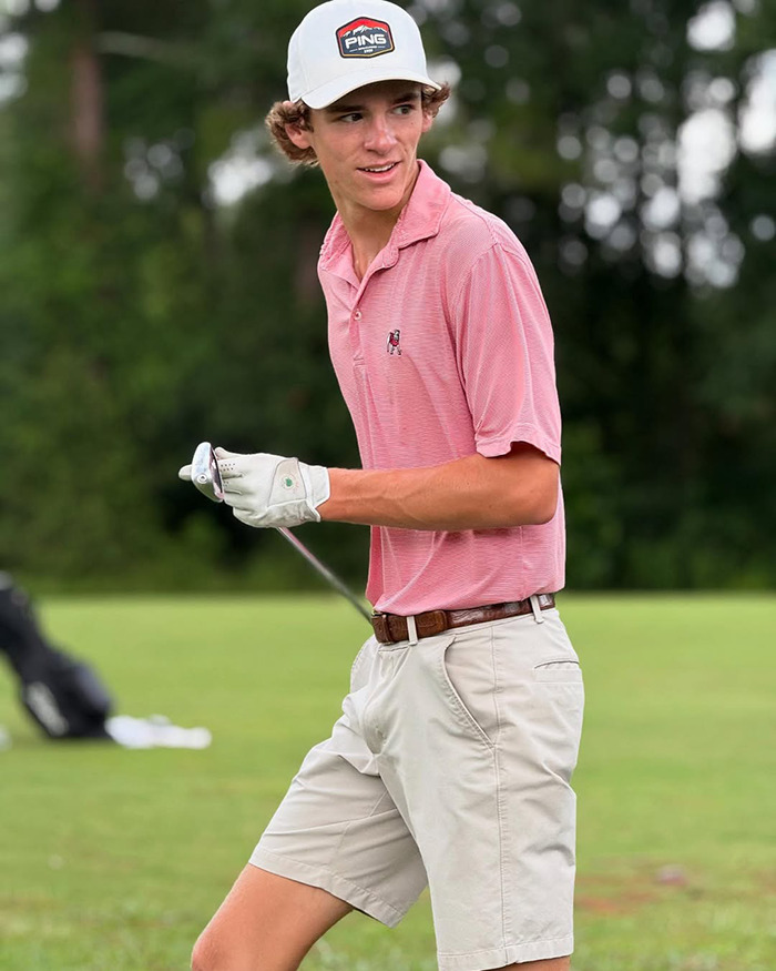 Teen golfer in pink polo and white cap holding a golf club on a green course during a practice session.