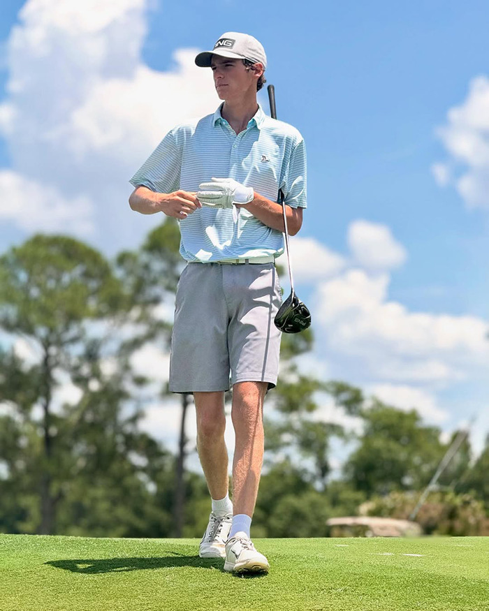 Promising teen golfer standing on green with club, dressed in light blue polo and gray shorts on sunny day.
