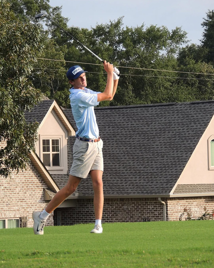 Teen golfer in blue shirt and cap swinging club on green lawn near residential houses during practice.