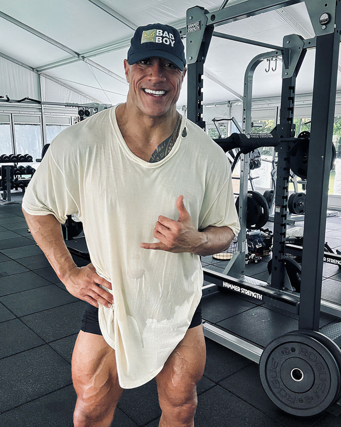 Dwayne Johnson in gym wearing a loose white shirt and black cap, showing his slim look after recent health concerns.