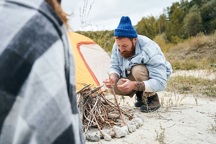 Man wearing a blue beanie building a campfire near a tent, illustrating things that look awesome in movies but are terrible in real life.