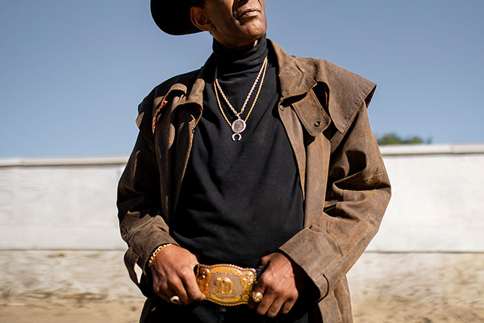 Man wearing a brown jacket and gold chains posing with a large belt buckle, illustrating things that look awesome in movies but are terrible in real life.
