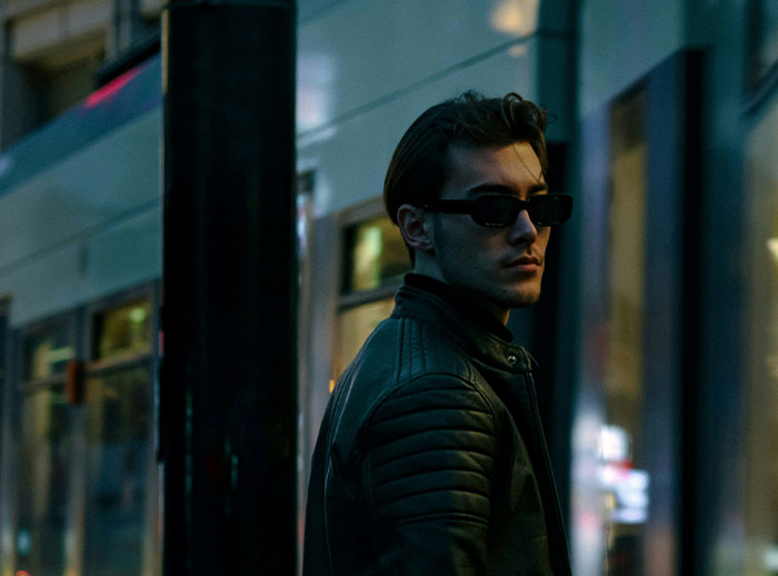 Man in dark sunglasses and leather jacket standing on city street at night representing things men do that affect women's safety.