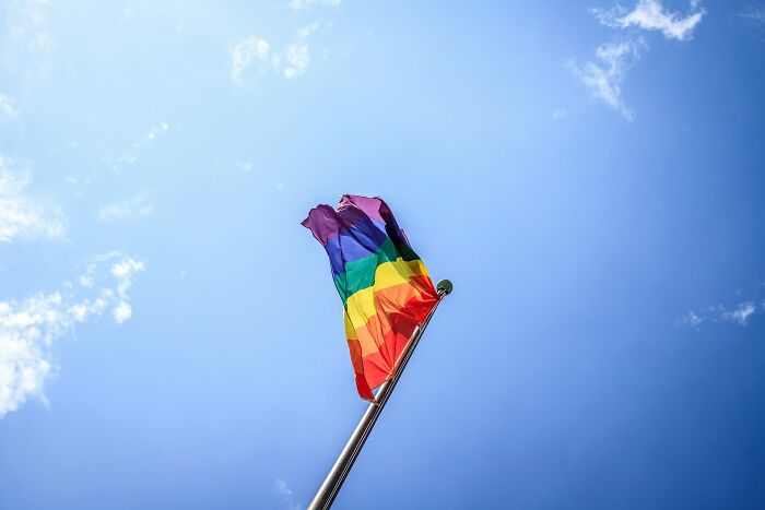 Colorful rainbow flag waving on a tall flagpole against a bright blue sky in a real life bizarre scene