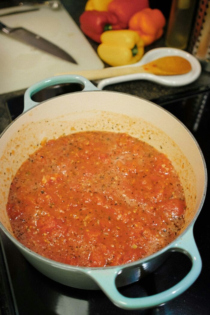 Simmering tomato sauce in a pot on the stove with fresh peppers and kitchen tools in the background for kitchen game tricks.