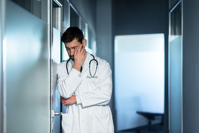 A doctor in a white coat and stethoscope leaning against a wall, reflecting on haunting medical experiences.