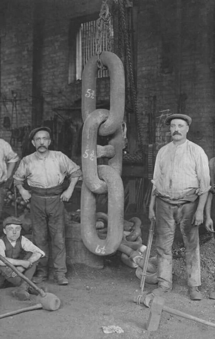 Black and white photo of workers posing with large heavy metal chains used in Titanic construction in a historic industrial setting.