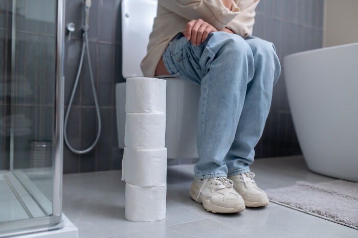 Person sitting on toilet in bathroom with stack of toilet paper rolls nearby, illustrating out-of-touch moments.
