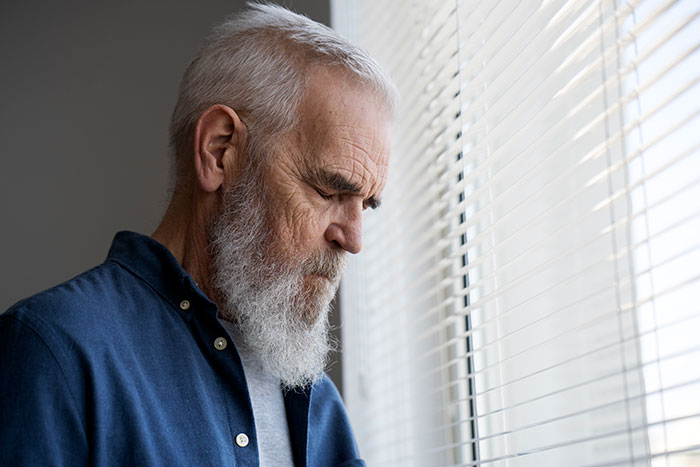 Elderly man with a white beard looking thoughtfully out a window, reflecting on lies that changed someone’s life.