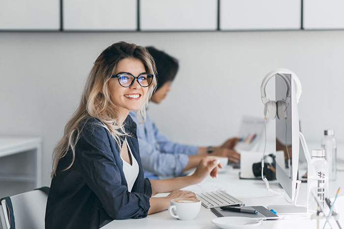 Young woman wearing glasses smiling at her desk with computer, illustrating lies that changed someone else’s life.