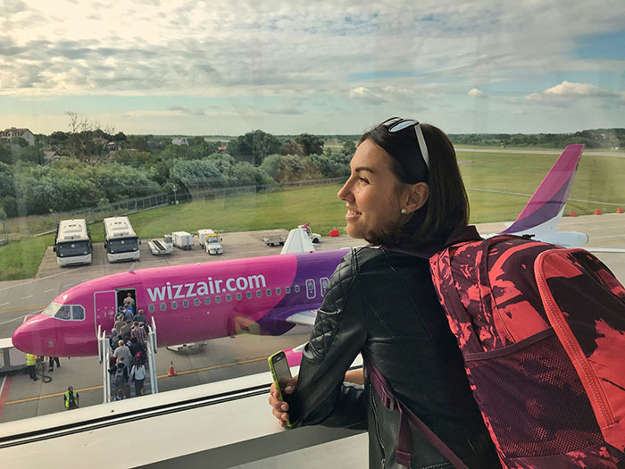 Young woman with a red backpack looking out at a Wizz Air plane, symbolizing life changes influenced by impactful lies.