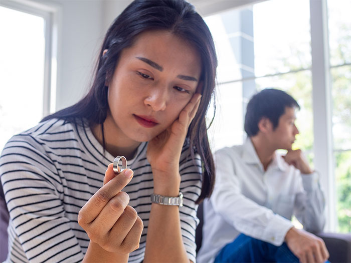 Woman holding a wedding ring looking upset while man sits distant in the background, hinting at cheating and divorce.