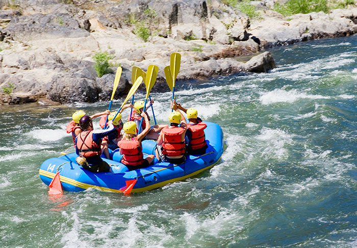 Group of tourists wearing life jackets and helmets rafting on rapid river water with paddles raised.