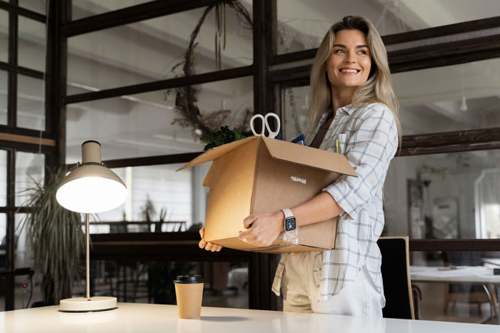 Smiling employee carrying a box with personal items, leaving office ahead of disciplinary hearing, surprising toxic bosses.