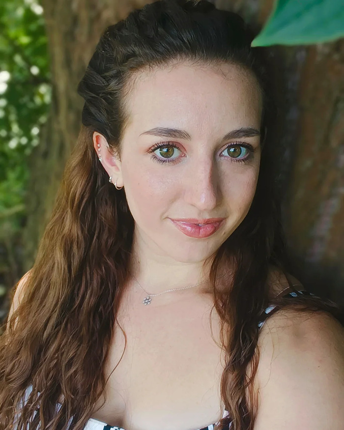 Young woman outdoors with long brown hair, wearing a necklace and striped top, reflecting on trapeze artist incident.