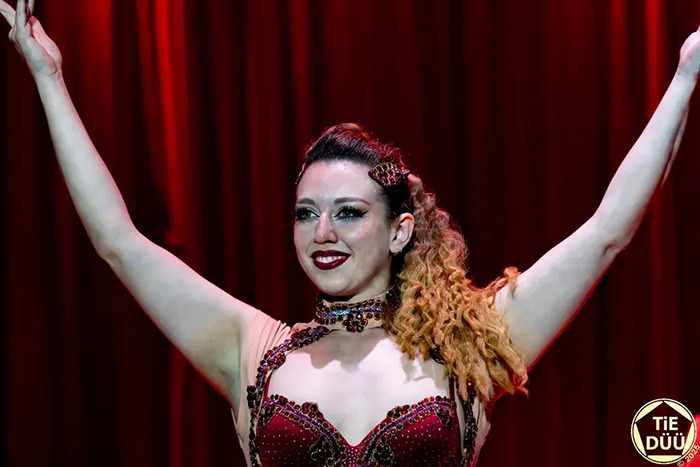 Trapeze artist in red costume with arms raised on stage, performing during a circus stunt in front of an audience.