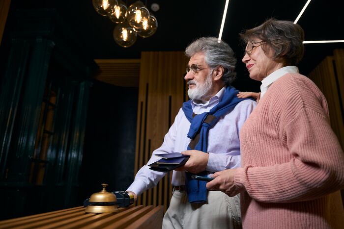 Elderly couple standing at hotel reception desk with bell, showcasing unexpected ways people died stories.