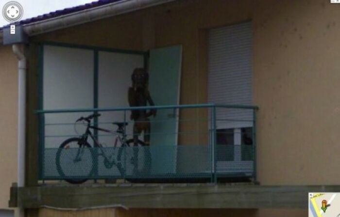 Bicycle parked on a balcony with a wooden statue figure behind a partially open door in strange unexplained photo.