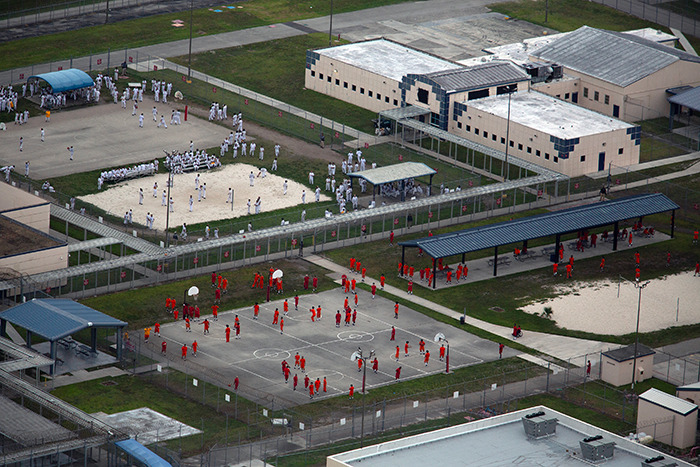 Aerial view of detainees in an ICE detention center yard with fences and multiple recreation areas visible.