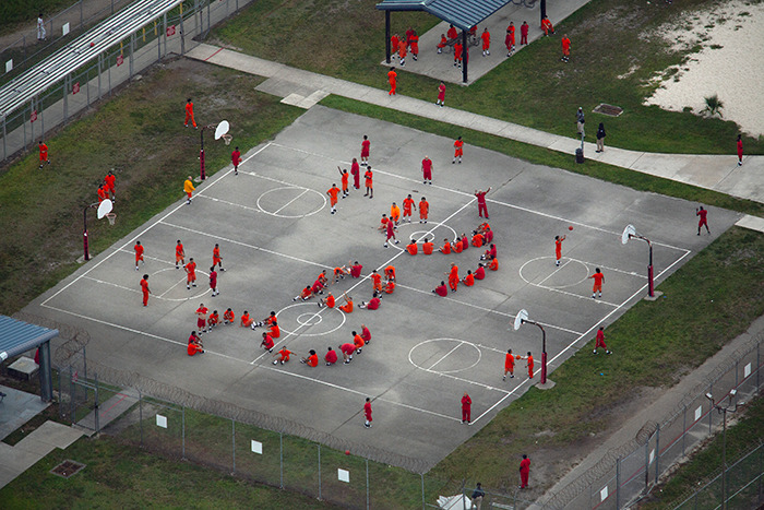 Aerial view of detainees in orange uniforms gathered on an outdoor basketball court at an ICE detention facility.