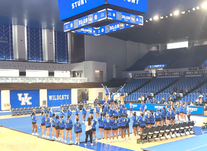 Cheerleader team performing stunts in a gymnasium with Kentucky Wildcats branding and blue uniforms. Cheerleader team performing stunts in a gymnasium with Kentucky Wildcats branding and blue uniforms.