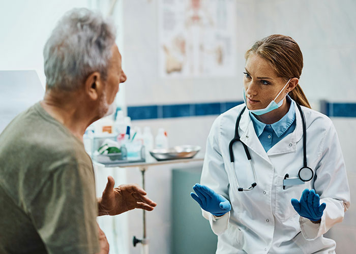 Doctor wearing gloves and mask talking attentively to senior patient in a medical office about industry secrets.