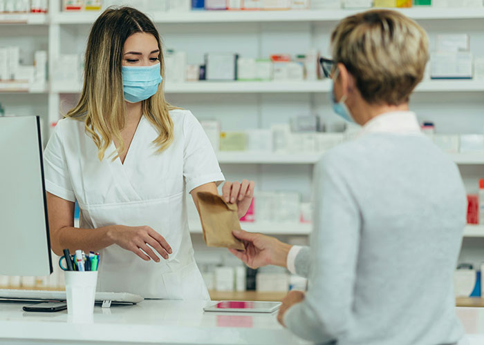 Pharmacist wearing a mask handing a brown paper bag to a customer in a modern pharmacy, revealing industry secrets.