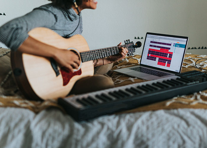 Person playing acoustic guitar next to a laptop and keyboard, exploring unexpected industry secrets in music production.