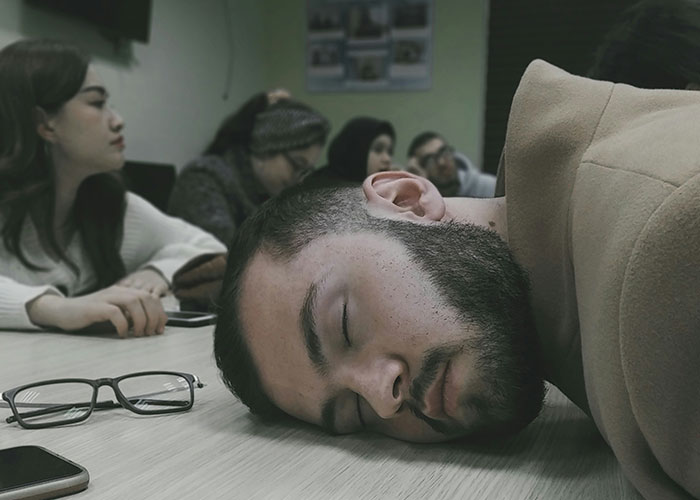 Man with beard sleeping on table during a meeting while coworkers sit in the background learning industry secrets.