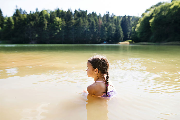 Young girl with braided hair standing in shallow lake water surrounded by trees, evoking paranormal and bizarre experiences.