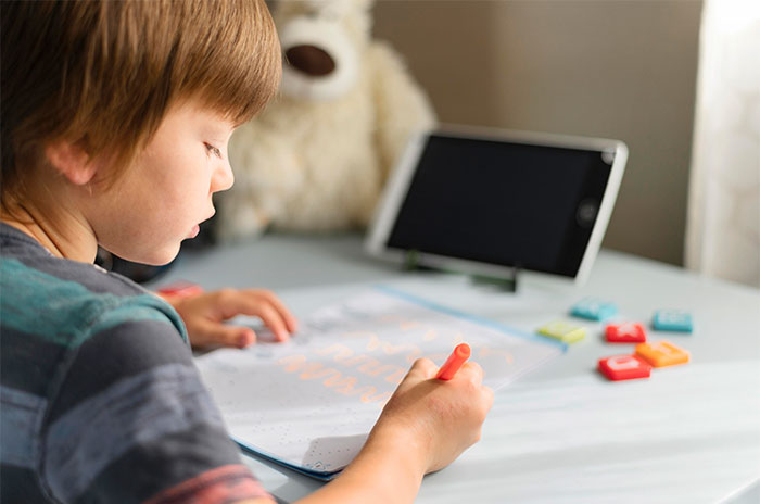 Young boy focused on a worksheet at a desk with a tablet and toys, illustrating bizarre and paranormal experiences concept.