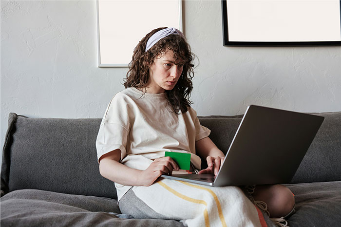 Young woman with curly hair and headband using laptop on sofa, focused on researching bizarre and paranormal experiences online.