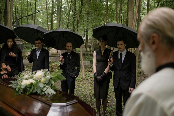 Mourners holding black umbrellas at a forest funeral, capturing a bizarre and paranormal atmosphere during the ceremony.