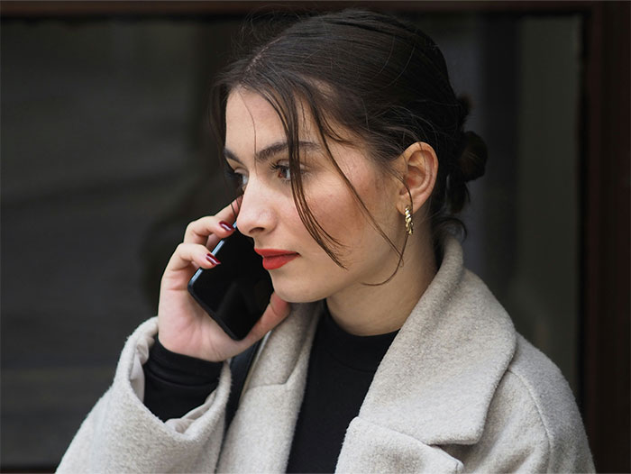 Young woman in a beige coat talking on a phone, reflecting on bizarre and paranormal experiences she can't explain.