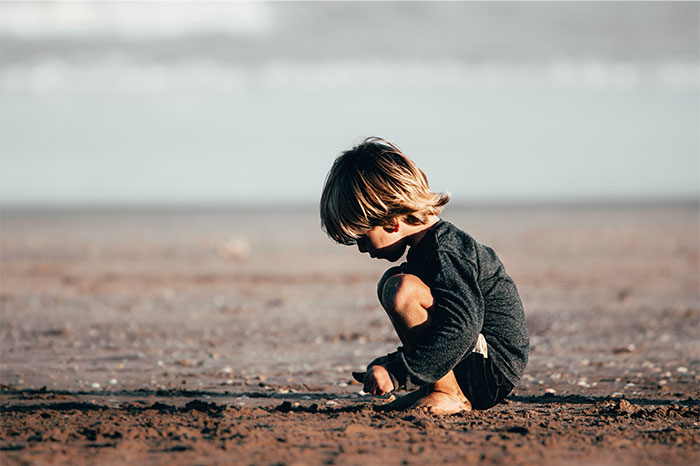 Young child crouching on sandy beach, focused and contemplative, evoking a sense of bizarre and paranormal experiences.