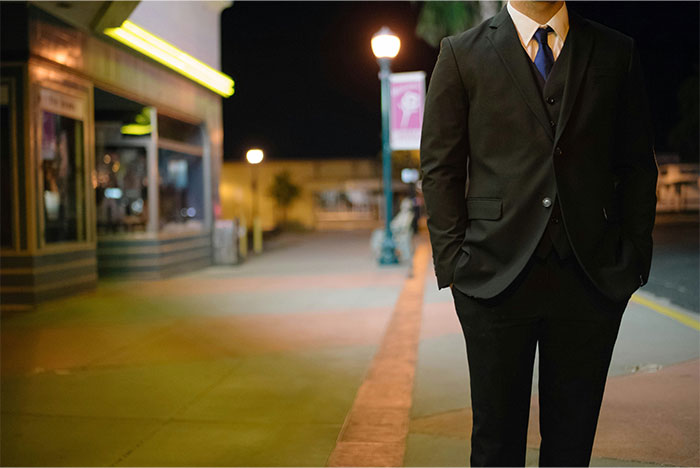 Man in a black suit standing on a sidewalk at night with blurred streetlights and buildings in the background, paranormal theme.