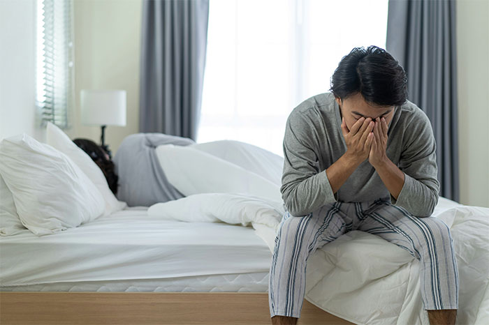 Man in pajamas sitting on bed with hands covering face while another person lies in bed, depicting bizarre paranormal experience