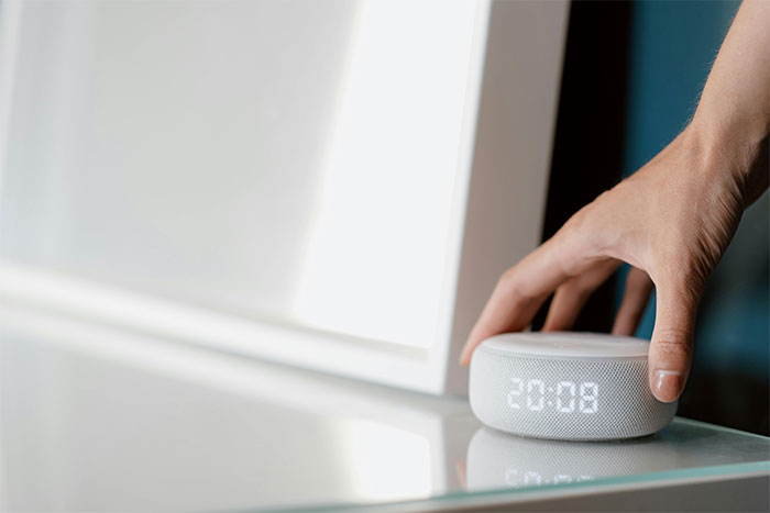 Hand reaching to press a smart speaker clock on a glass table, evoking strange and paranormal experiences concept.