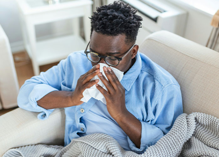 Man sitting on a couch sneezing into a tissue, illustrating unusual body quirks and fascinating human body reactions.