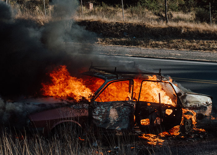 Burning car engulfed in flames and thick smoke near a roadside, portraying a nightmare vacation gone terribly wrong.