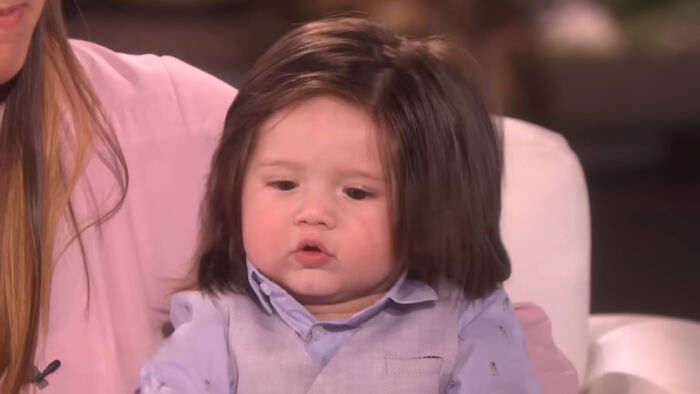 Baby boy with thick dark hair wearing a blue shirt and vest sitting on a white chair, related to shocking school incidents.