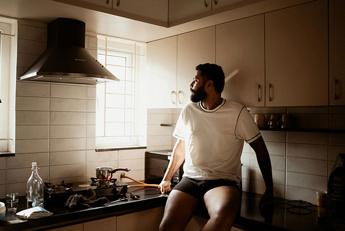 Man in boxers sitting on kitchen counter by window, with natural light highlighting the awkward moment involving boxers and guests.