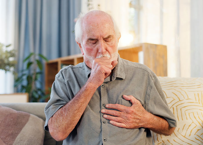 Elderly man coughing into fist while sitting on couch, illustrating wild rules people grew up with health concerns.