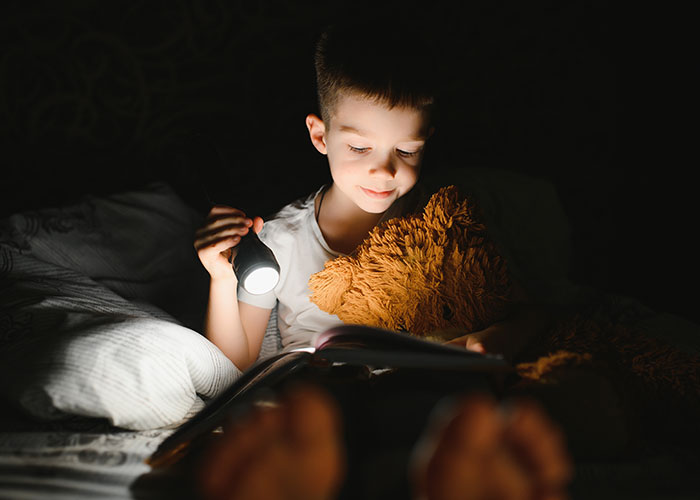 Young child holding a flashlight and reading a book at night, reflecting on wildest rules people grew up with.
