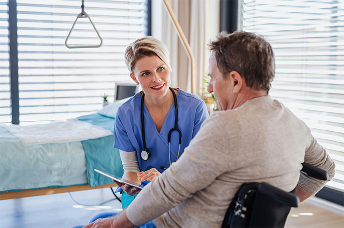 Female doctor in blue scrubs talking to elderly male patient in wheelchair during medical consultation.